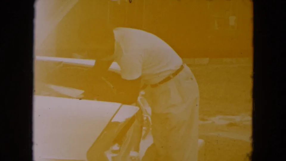 The man remains bent over the open trunk of the light-colored classic car, continuing to load luggage in the home-movie footage.