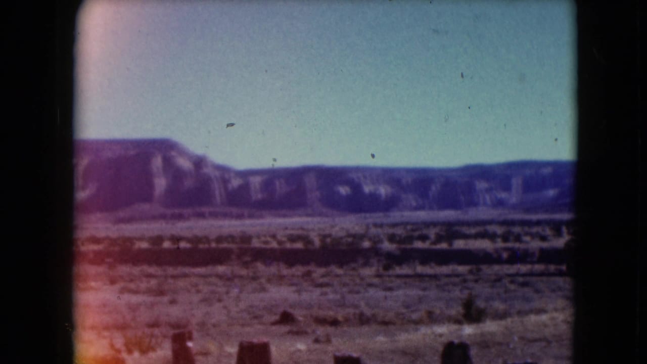 1961 Nevada 8mm frame showing a sandy desert plain, low shrubs, distant purple-toned mountains, and a hazy sky.