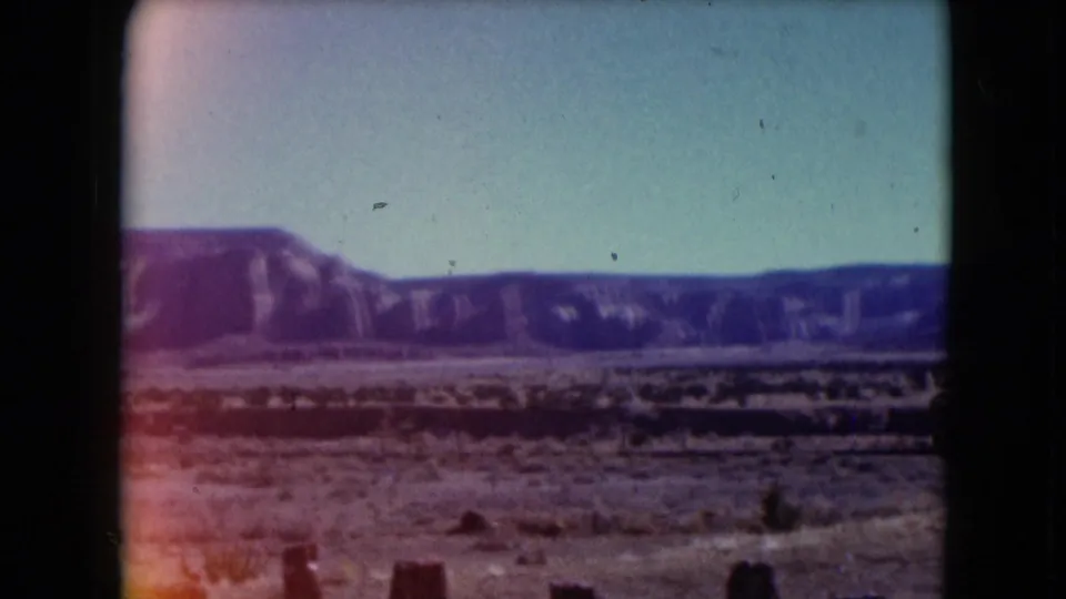 1961 Nevada 8mm frame showing a sandy desert plain, low shrubs, distant purple-toned mountains, and a hazy sky.