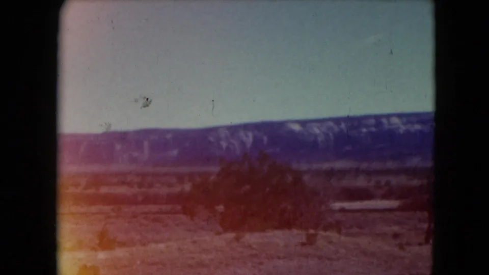 Faded 1961 Nevada 8mm frame showing desert shrubs, sandy ground, distant mountains, and a pale blue-green sky.