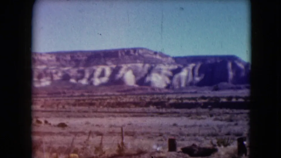 1961 Nevada 8mm frame showing dry desert scrub, roadside posts, and a broad rocky escarpment beneath a clear sky.