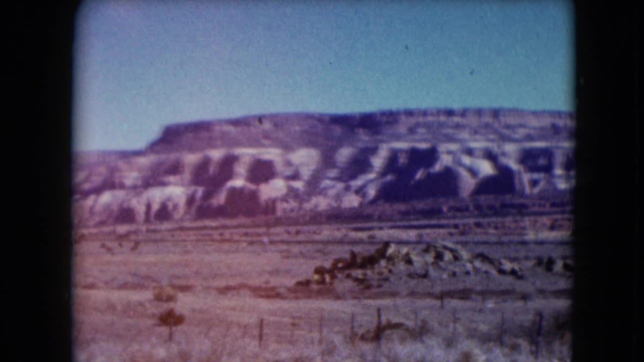 1961 Nevada 8mm frame showing a broad desert mesa, pale eroded rock, sparse brush, and a low wire fence in the foreground.