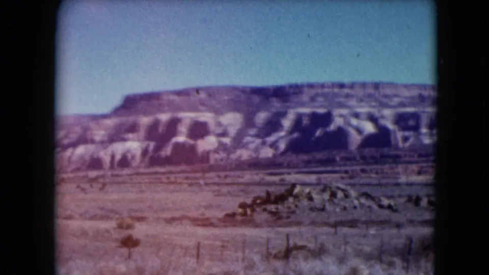 1961 Nevada 8mm frame showing a broad desert mesa, pale eroded rock, sparse brush, and a low wire fence in the foreground.