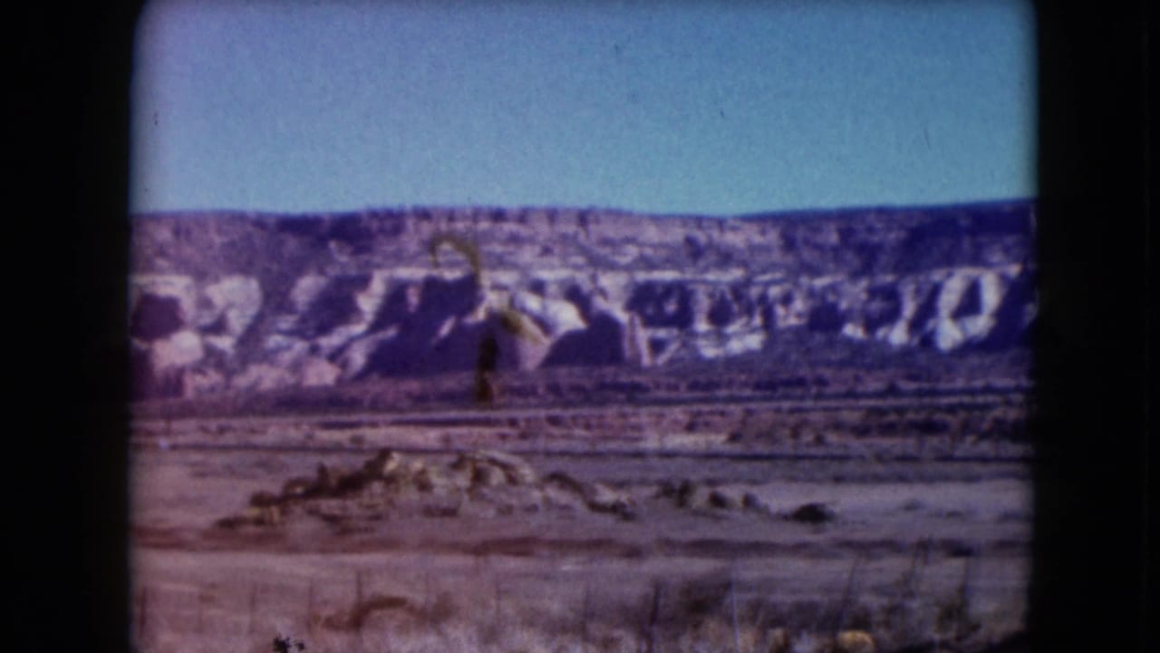 Wide desert landscape with mesa cliffs, scrub, and fence posts in the foreground.