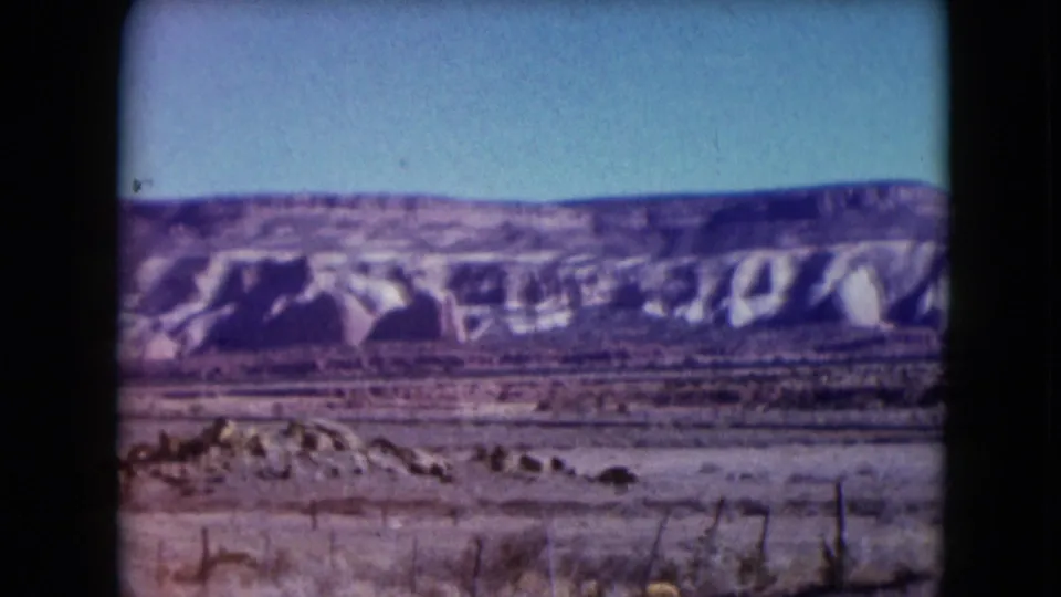 1961 Nevada 8mm frame with the mesa shifted across the view, showing eroded formations, low scrub, fence lines, and a clear sky.