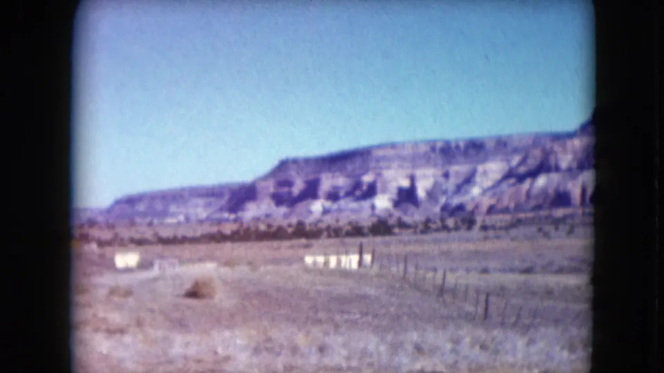 1961 Nevada desert view with pale open ground, fence posts, scrub, and eroded hills under a blue sky.