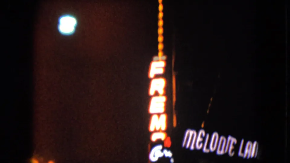 1961 Nevada home-movie frame with a vertical Fremont neon sign and Melodie Lane lettering against mostly dark surroundings.