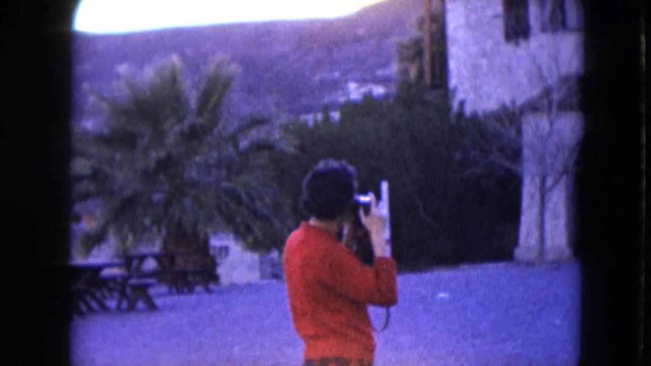 1961 Nevada 8mm frame showing a person in a red sweater using a camera near picnic tables, palm trees, a stone structure, and arid hills.