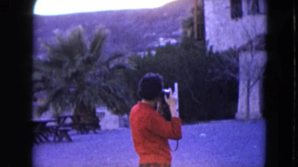 1961 Nevada 8mm frame showing a person in a red sweater using a camera near picnic tables, palm trees, a stone structure, and arid hills.