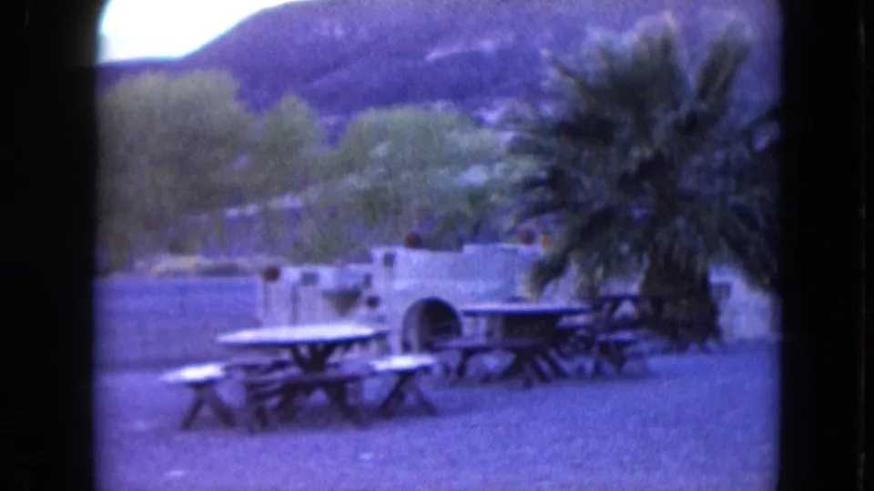 1961 Nevada home-movie frame of an open picnic area with tables, palm trees, low stonework, and mountains beyond.