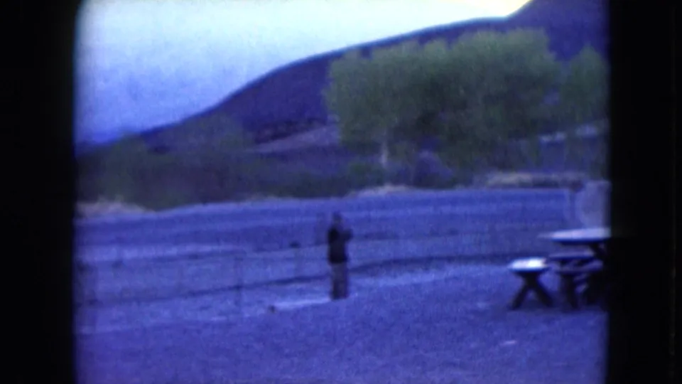 1961 Nevada 8mm frame with a small distant person near fencing, picnic tables, open gravel ground, trees, and low mountains.