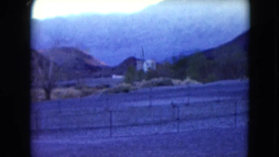 1961 Nevada home-movie frame showing fencing, brush, a palm tree, open ground, and layered desert mountains in soft color.
