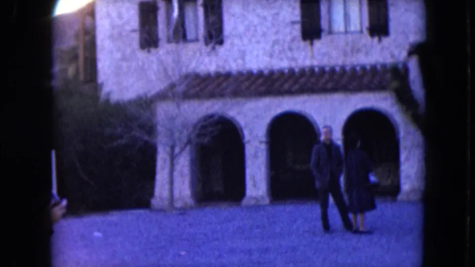 Home-movie frame of two adults crossing the paved foreground before an arched stone exterior in Nevada, 1961.