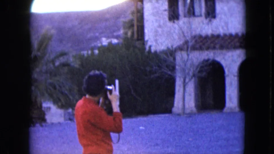 A red-sweater visitor photographs the historic-style exterior, with arches, landscaping, and distant hillside visible.