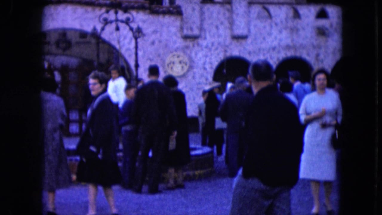 People stand and walk in front of arched stone doorways, with a woman in a light dress at right.