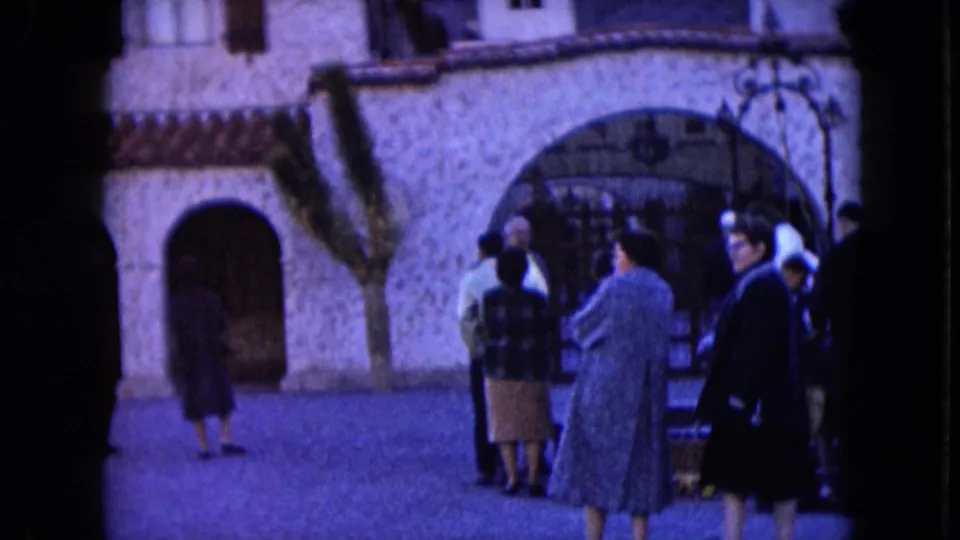 Wide 1961 Nevada home-movie view of adults outside a stone building with arched openings and a small door.