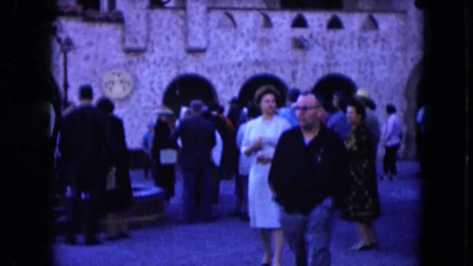 Home-movie frame from Nevada in 1961 with adults passing the camera and visitors gathered near stone arches.
