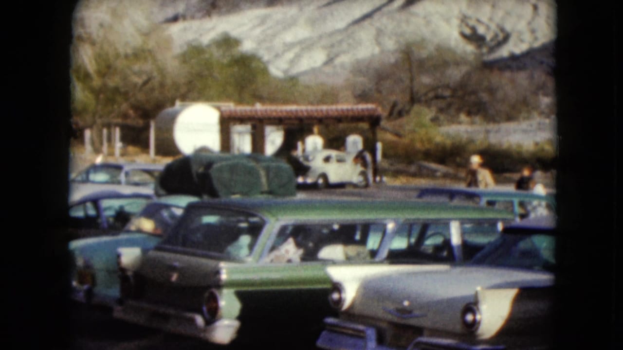 1961 Nevada home-movie frame showing parked American cars near a rustic roadside shelter, with distant people and hills behind.