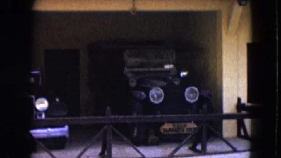 A handheld 8mm view shows antique cars behind a railing, including a dark vehicle with prominent round headlights.