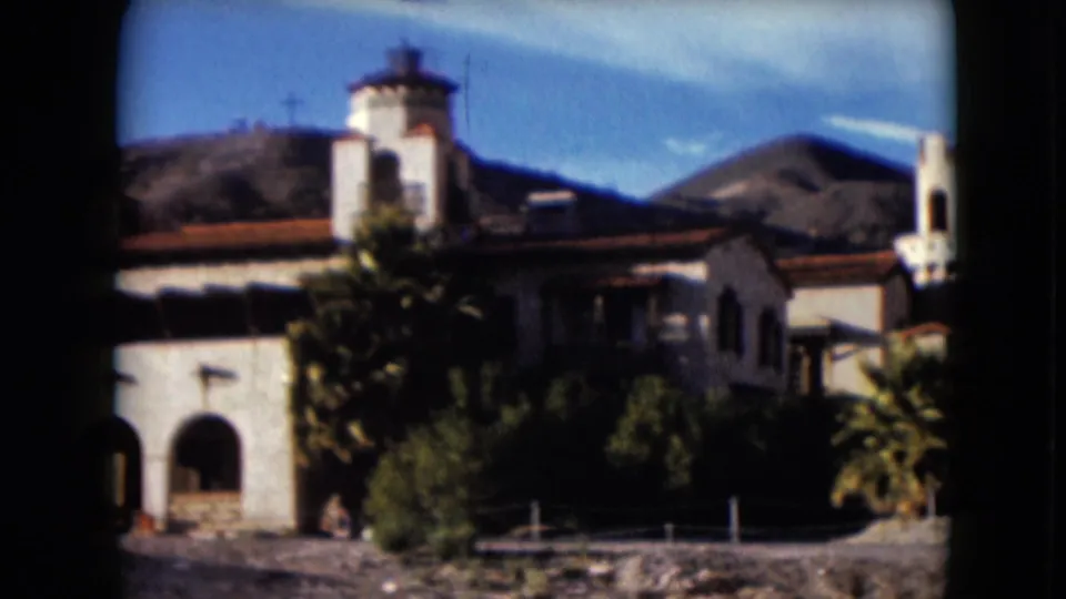 1961 Nevada 8mm frame showing a stucco Spanish-style building with arched entryways, tower, palms, and rocky mountains behind it.