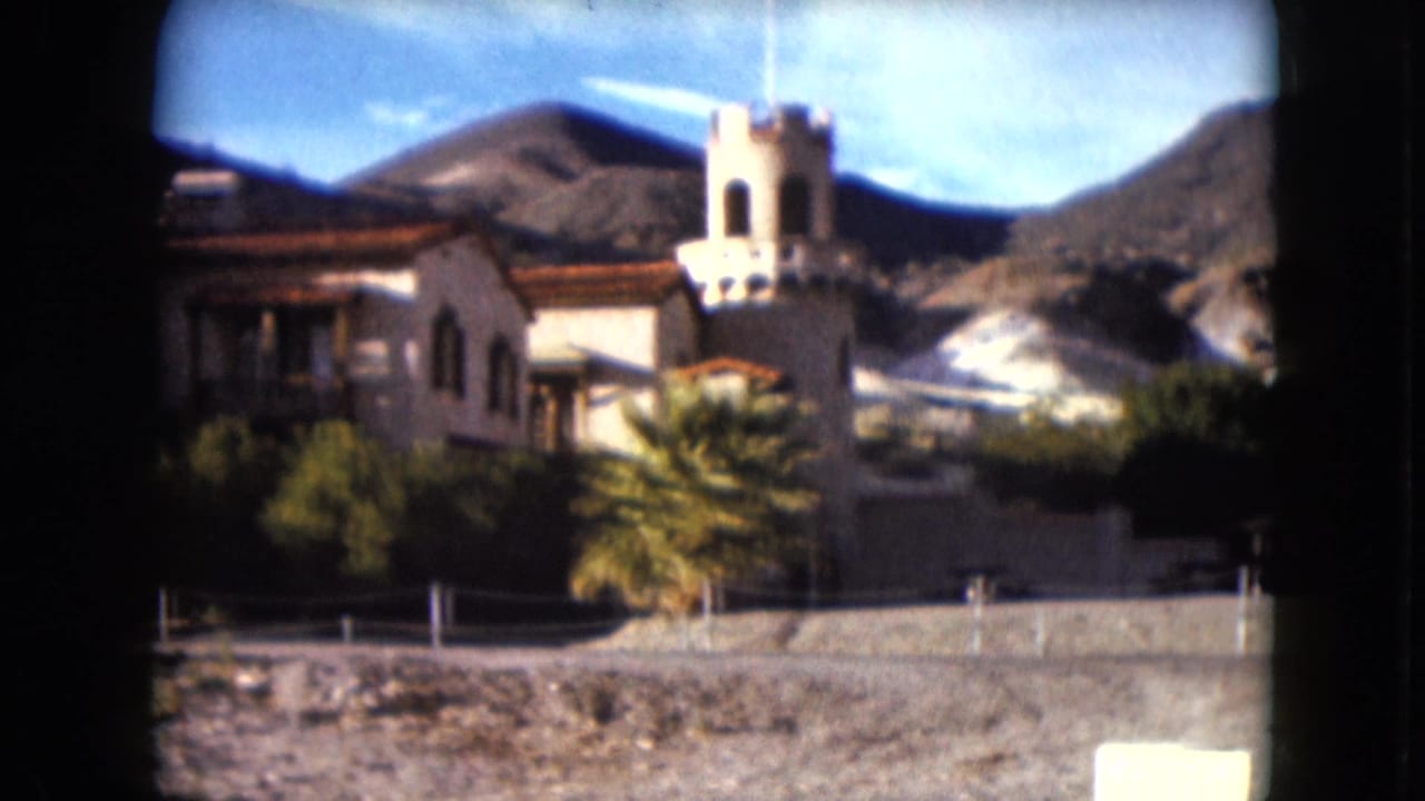 1961 Nevada frame of a Spanish-style building and prominent tower, seen across a low wall with palms and desert hills behind.