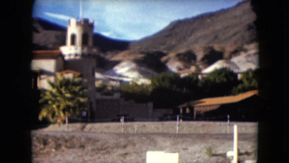 1961 Nevada home-movie view of the building's tower, palms, low fencing, and pale rocky slopes beyond the courtyard area.