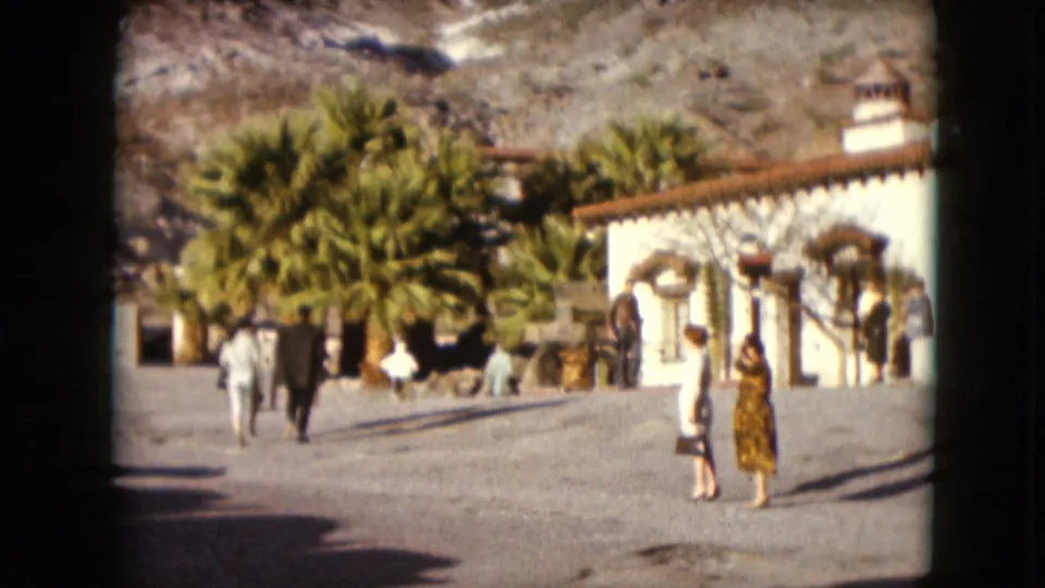 1961 Nevada frame with several distant visitors walking through a courtyard beside palms, stucco walls, arched windows, and desert hills.