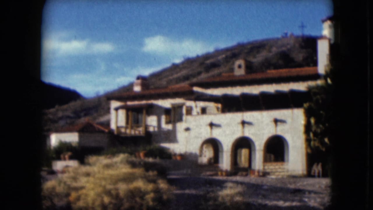 Stucco building with arched openings, red roof, shrubs, and hillside.