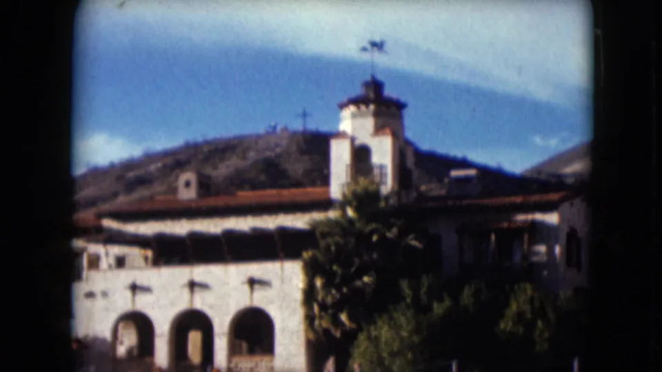 1961 Nevada frame with a Spanish-style stucco building, arched openings, tower, trees, hillside, and a cross visible on the ridge.