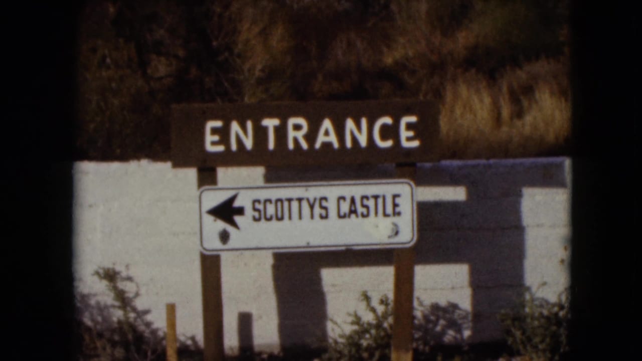 1961 Nevada frame showing a brown ENTRANCE sign over a white left-arrow SCOTTYS CASTLE sign near a stucco wall and dry brush.