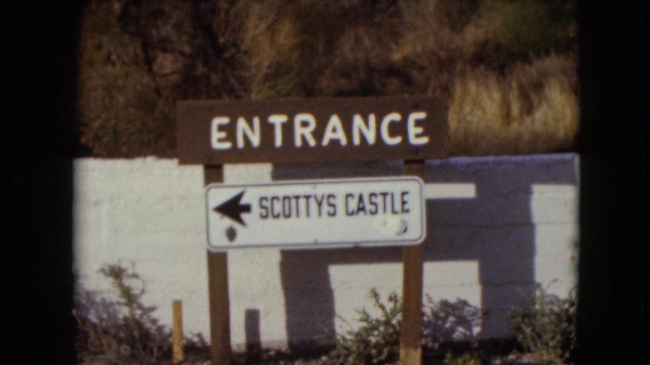 Brown ENTRANCE board and white SCOTTYS CASTLE arrow sign mounted on posts near a white wall.