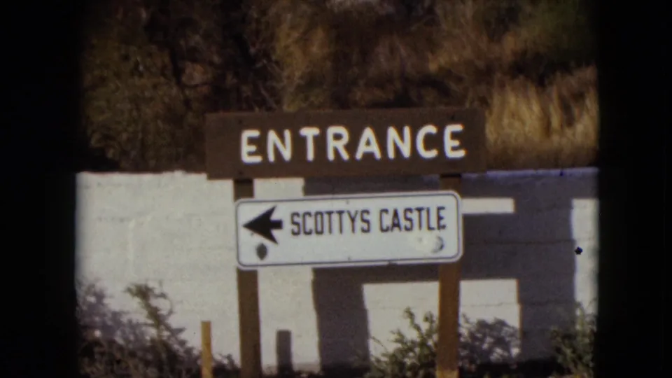 1961 Nevada frame with the Scotty's Castle entrance sign filling the shot, showing white lettering, a left arrow, posts, wall, and desert brush.