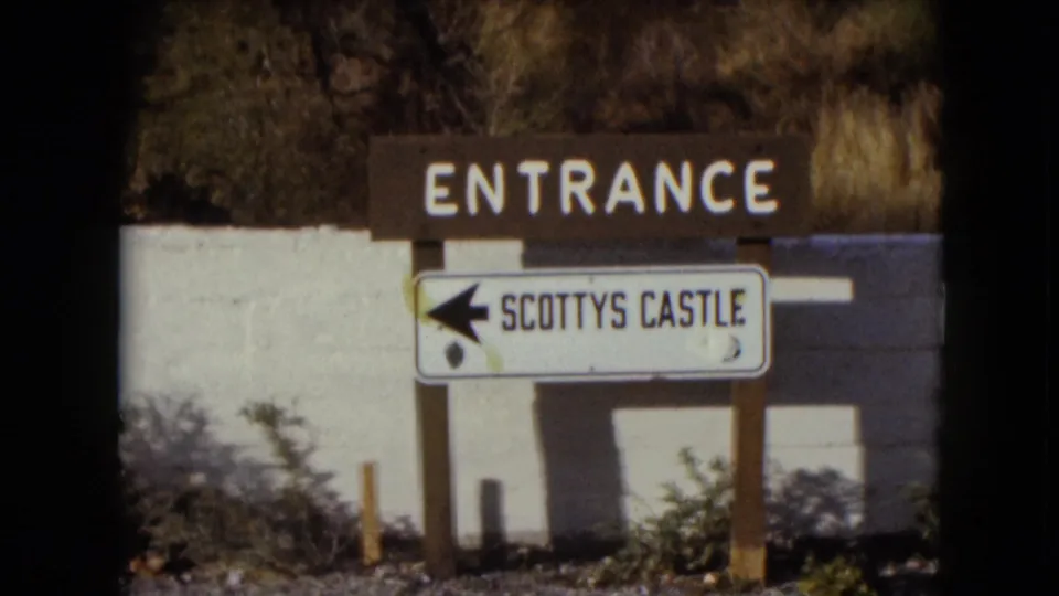 1961 Nevada home-movie frame showing the Scotty's Castle entrance wayfinding sign against a white wall with brush and strong sunlight.