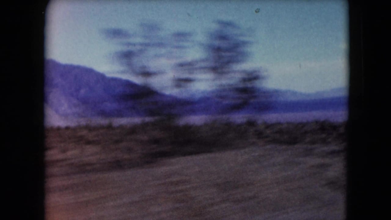 1961 Nevada 8mm road-view frame with blurred desert vegetation, sandy ground and distant mountains.