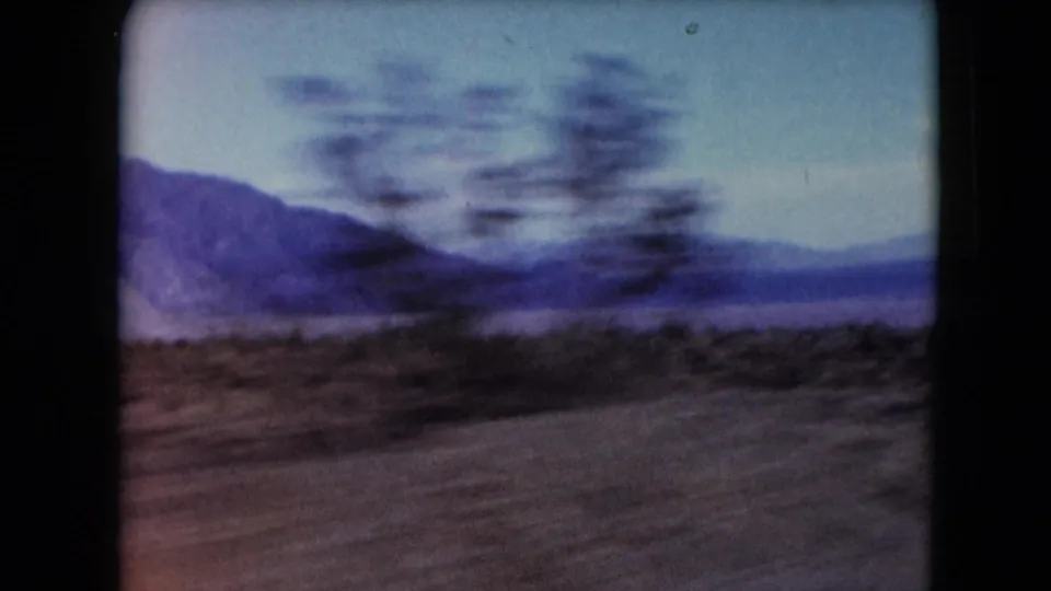 1961 Nevada 8mm road-view frame with blurred desert vegetation, sandy ground and distant mountains.