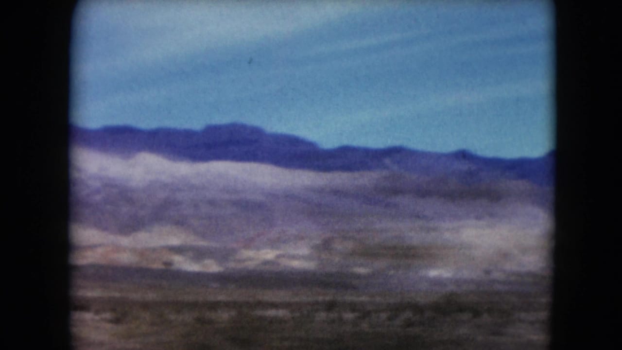 1961 Nevada 8mm frame showing distant desert mountains, open blue sky, and sparse arid foreground from a moving viewpoint.