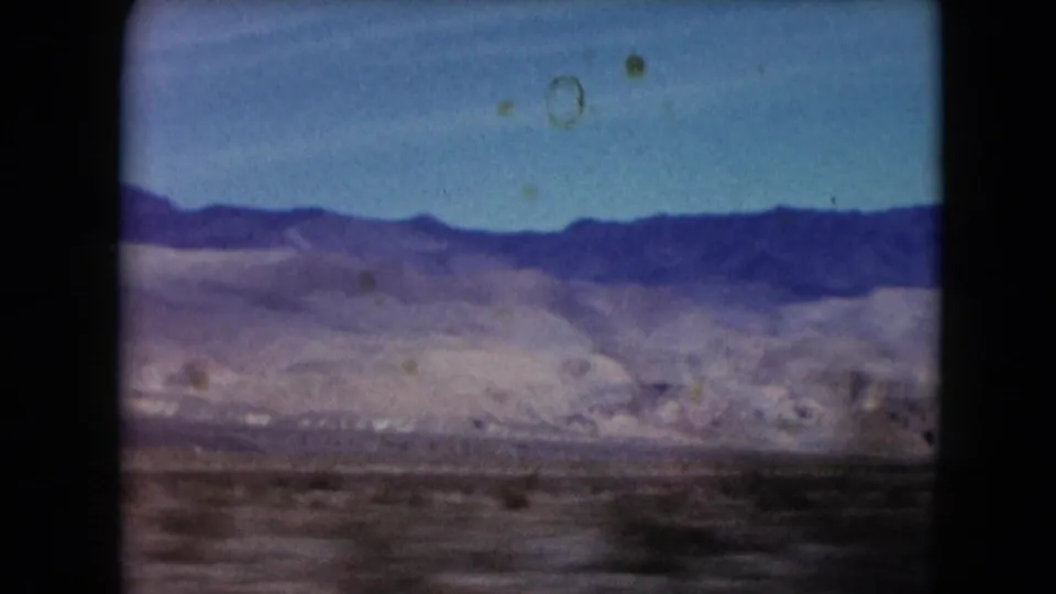 1961 Nevada home-movie frame with a dark mountain ridge, pale desert foothills, blue sky, and visible film dust.