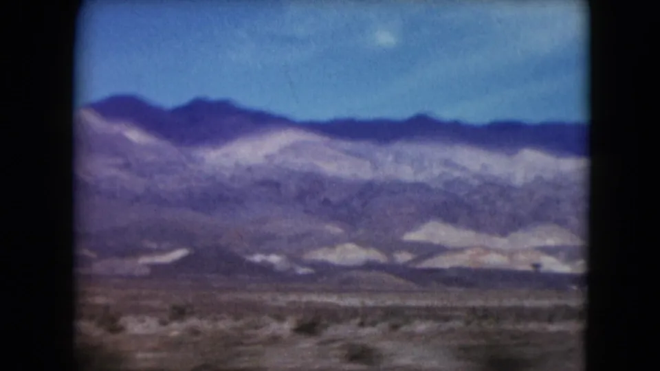 Nevada 1961 8mm frame showing pale mountain slopes, darker ridge line, and low desert vegetation from a roadside view.