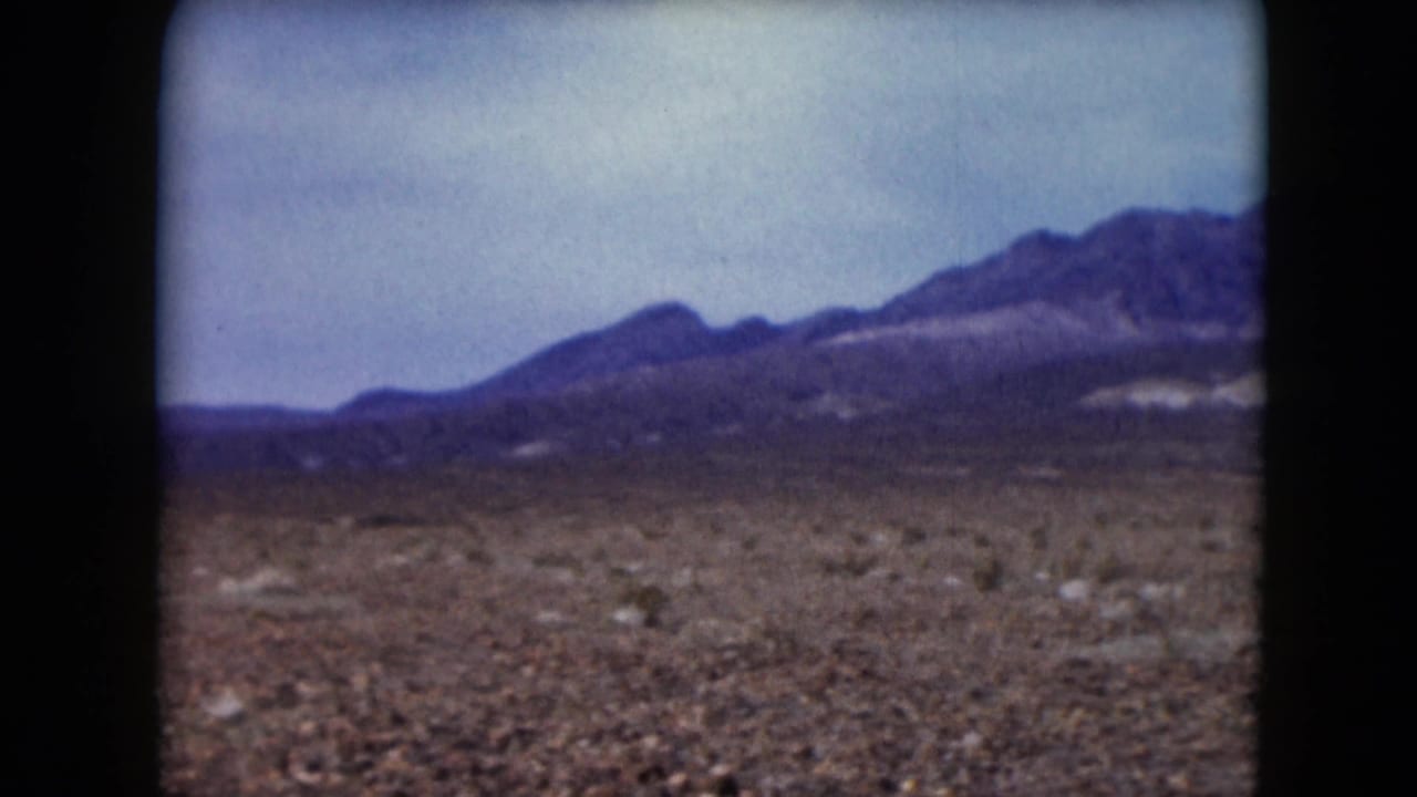1961 Nevada home-movie frame showing a desert plain, low brush, and distant mountain ridges beneath a cloudy sky.