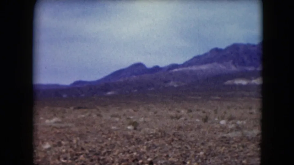 1961 Nevada home-movie frame showing a desert plain, low brush, and distant mountain ridges beneath a cloudy sky.