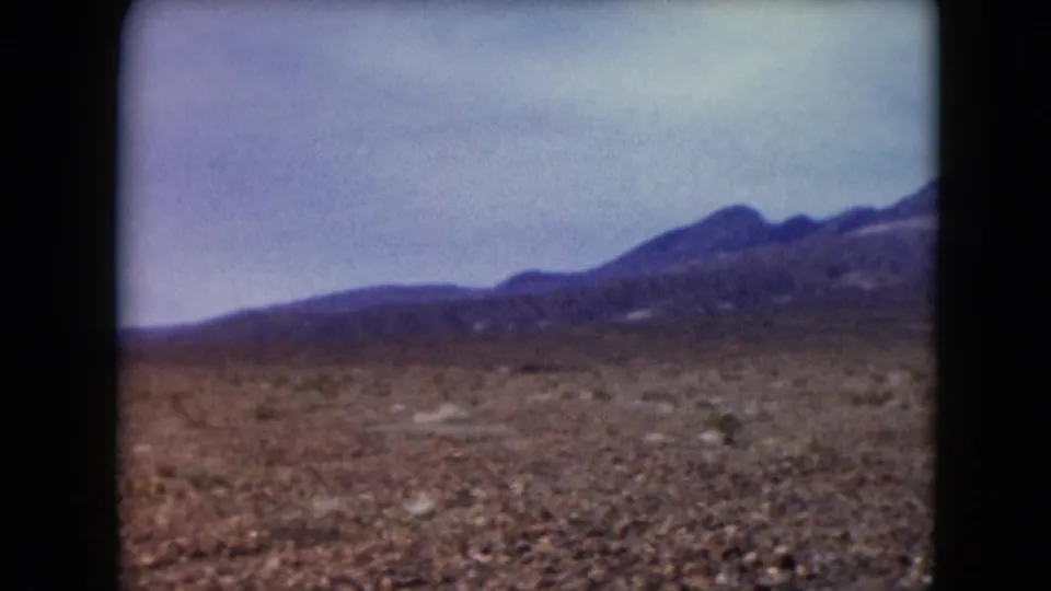 1961 Nevada desert view from a moving vehicle, with rocky ground, scattered shrubs, and distant mountain slopes under cloud cover.