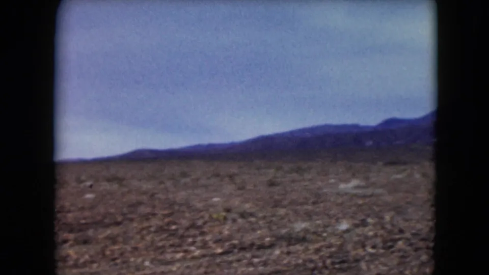 1961 Nevada landscape frame showing flat desert ground, scattered brush, and a low mountain range beneath a pale cloudy sky.