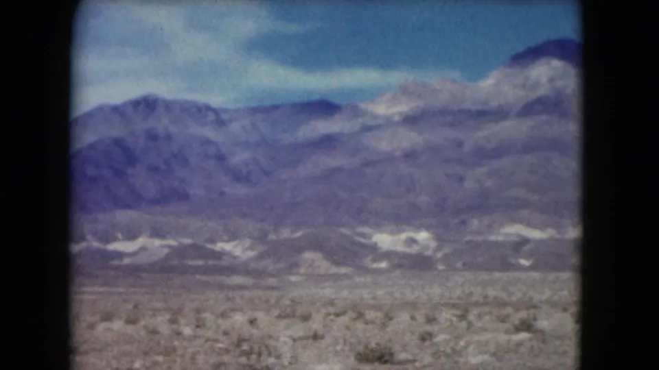 1961 Nevada home-movie frame showing a dry desert plain, scattered shrubs, and layered mountain ridges beneath a pale sky.