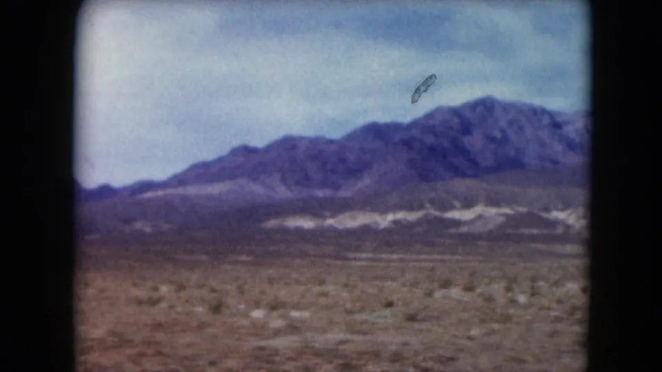 1961 Nevada landscape frame with a broad desert foreground, mountain ridge, pale sky, and a small dark speck overhead.