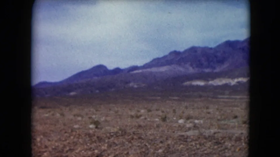 1961 Nevada home-movie frame showing rocky dry ground, sparse desert vegetation, and a distant mountain range.