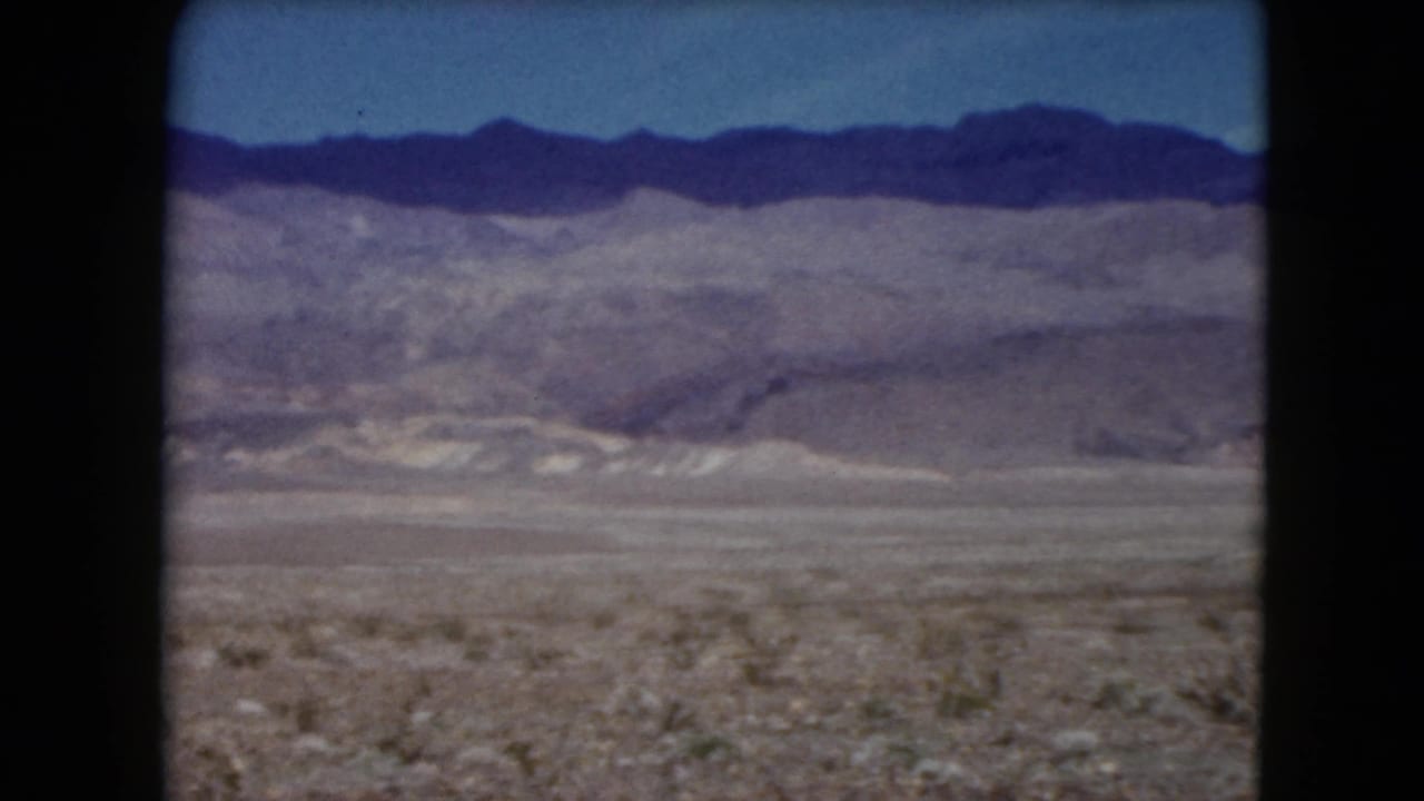 1961 Nevada 8mm frame showing a rocky desert plain with low brush and distant layered mountains under a clear blue sky.