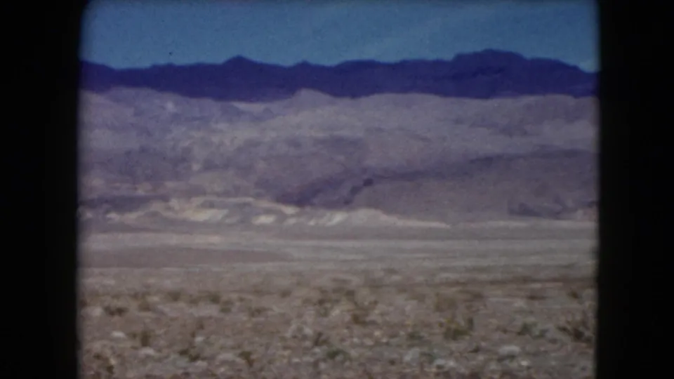 1961 Nevada 8mm frame showing a rocky desert plain with low brush and distant layered mountains under a clear blue sky.