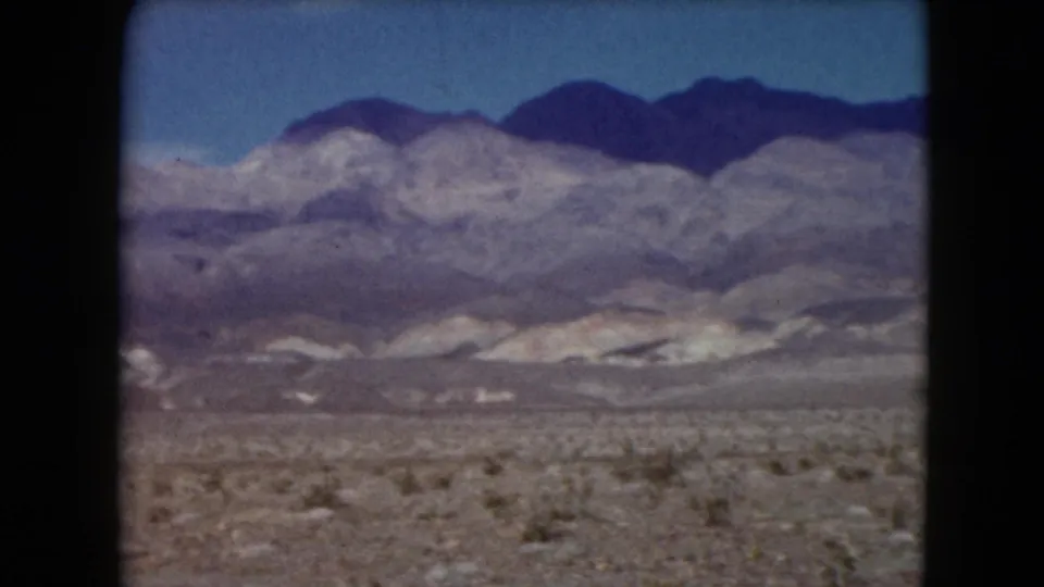 Nevada 1961 8mm frame showing low desert vegetation, open arid ground, and broad gray-purple mountain slopes.