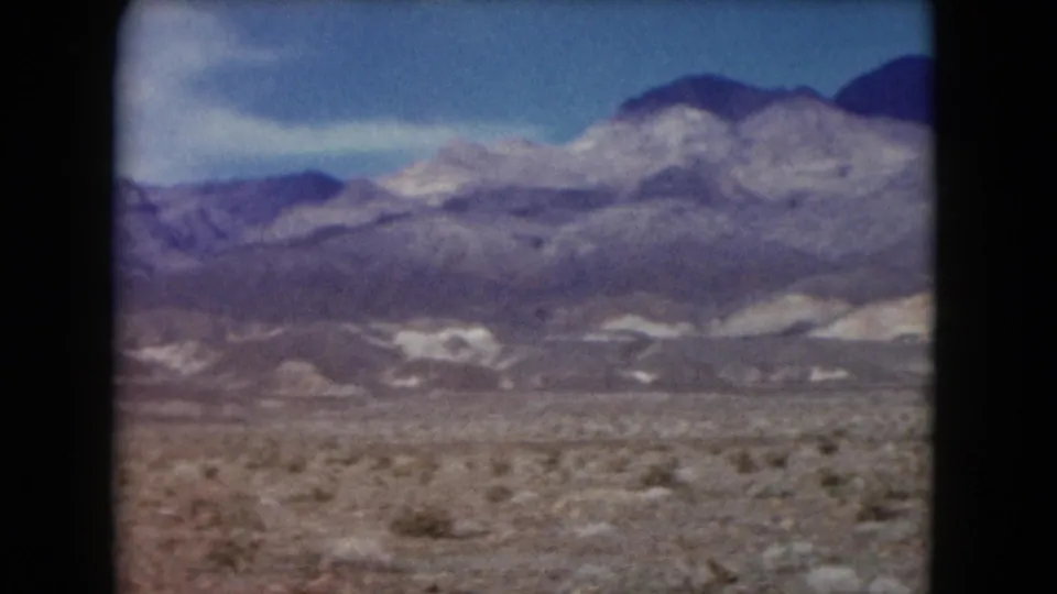 1961 Nevada 8mm still of desert scrub, layered mountains with pale patches, and blue sky with a small cloud.