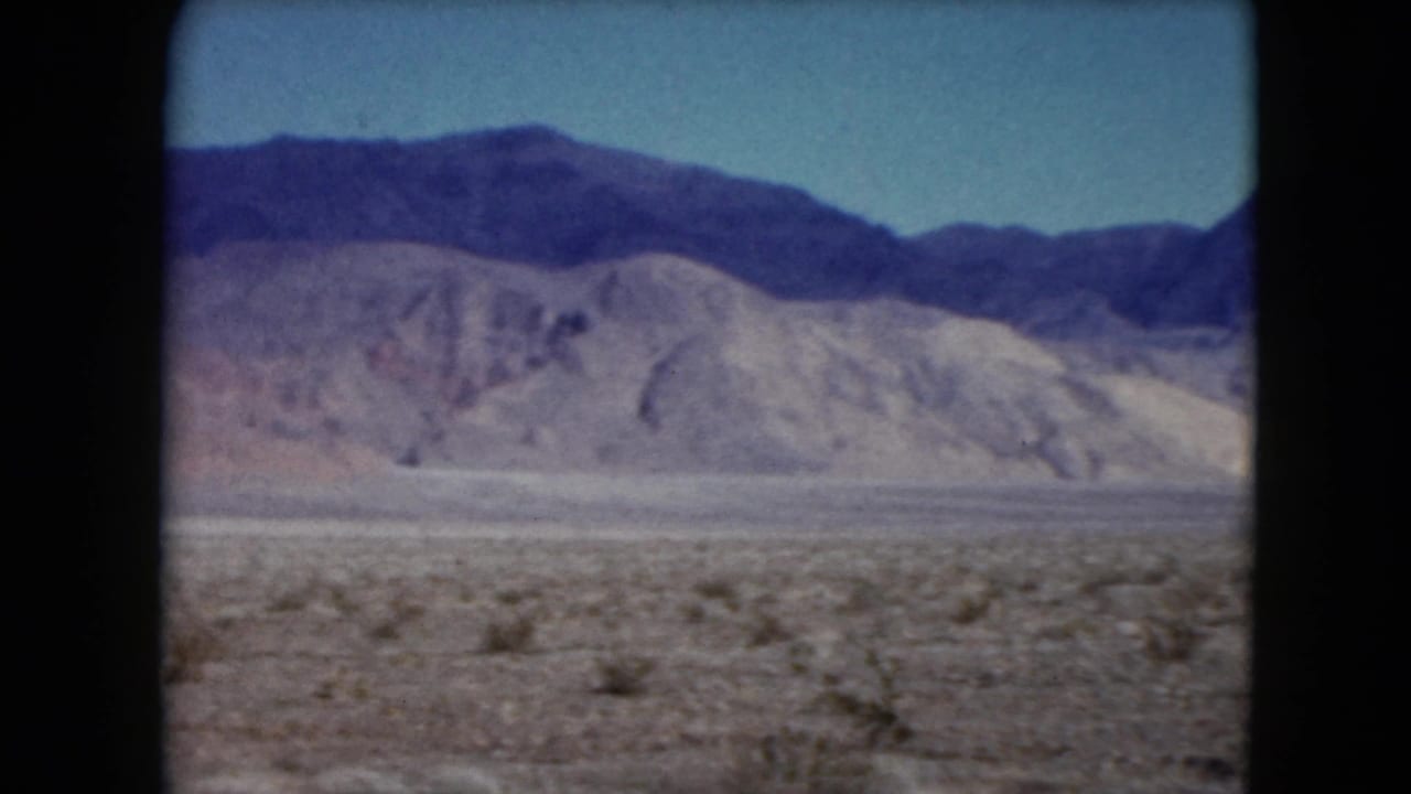 Desert plain with low scrub and distant mountains under a blue sky.